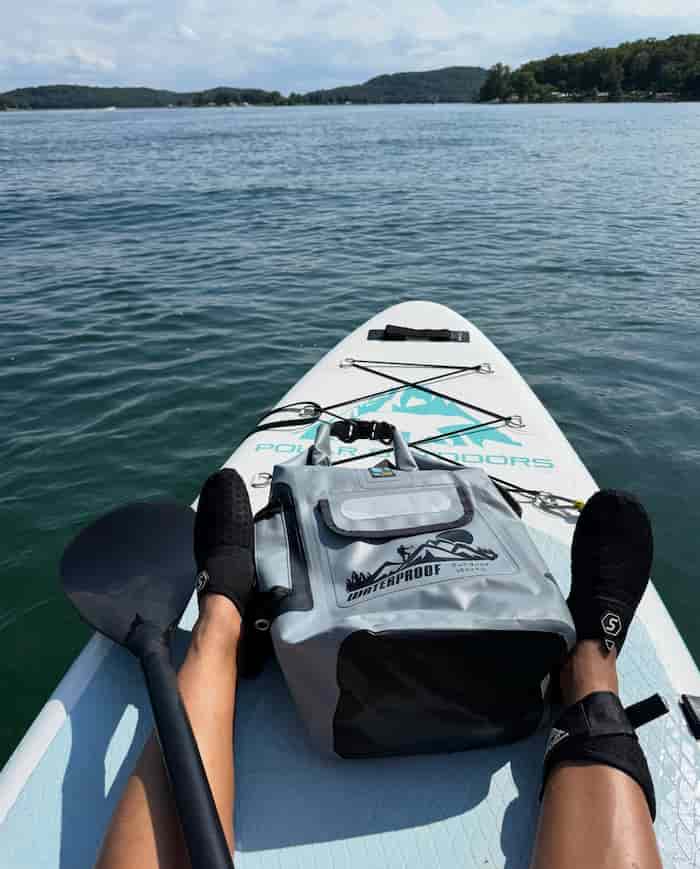 A person sitting on a stand-up paddleboard on open water, with an idrybag pro shield gray waterproof bag placed on the board in front of them. The board has teal branding text, and the person is wearing black foot covers.