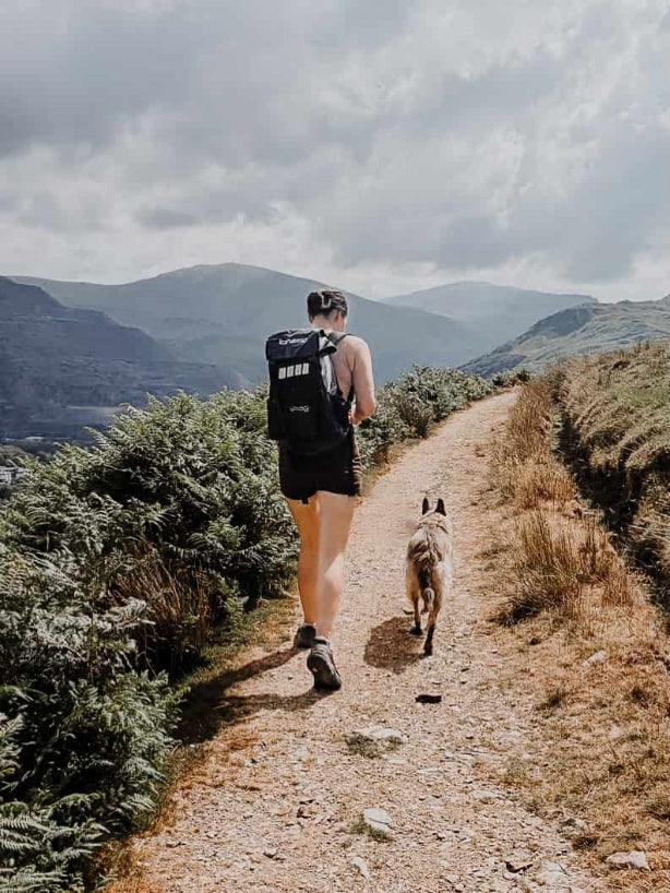 A woman hiking on a mountain trail with a dog, wearing an idrybag expedition voyager black backpack