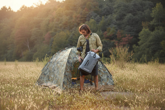 Person standing next to a camouflage tent in a forested area, holding a gray idrybag backpack and camera.