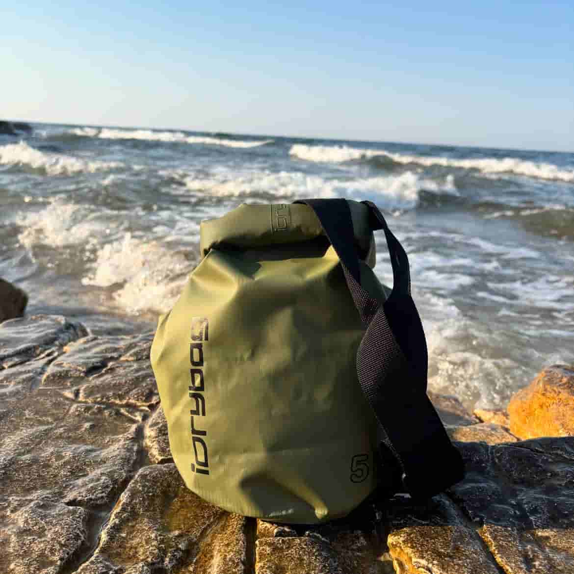 An olive green dry bag with a black strap sits on rocks with the ocean waves in the background. The brand name idrybag is printed vertically on the side.
