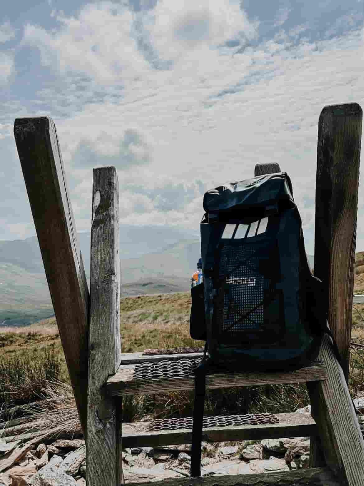 A black idrybag expedition voyager backpack sits on a weathered wooden bench in an outdoor mountain setting under a cloudy sky.