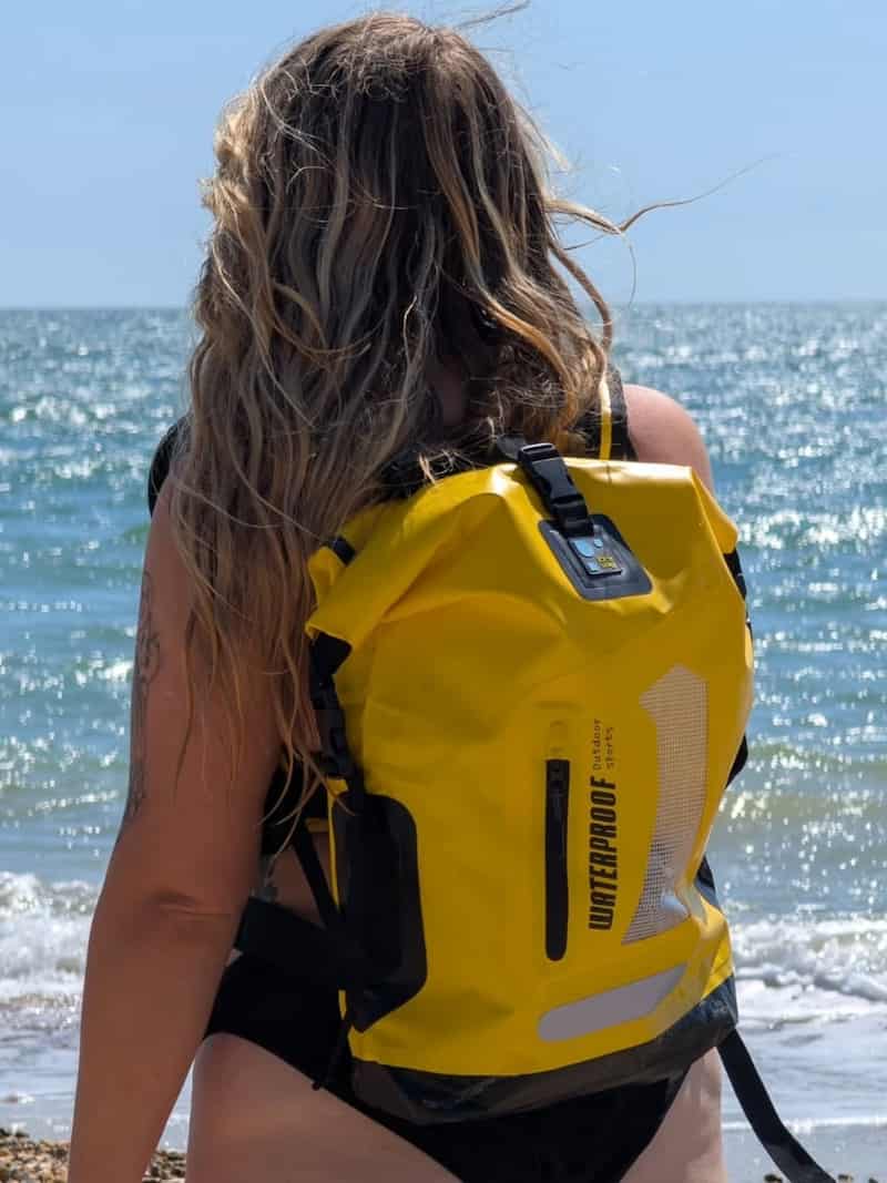 A woman seen from behind on a beach wearing a bright yellow idrybag pro lite waterproof dry bag backpack with black straps and reflective details, with the ocean in the background.
