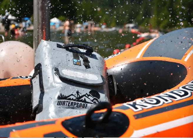 A close-up action shot of a grey waterproof backpack being splashed with water inside an orange and black inflatable raft. The raft has 'KONDOR' printed on it, and the background shows a blurry outdoor lake scene with people.