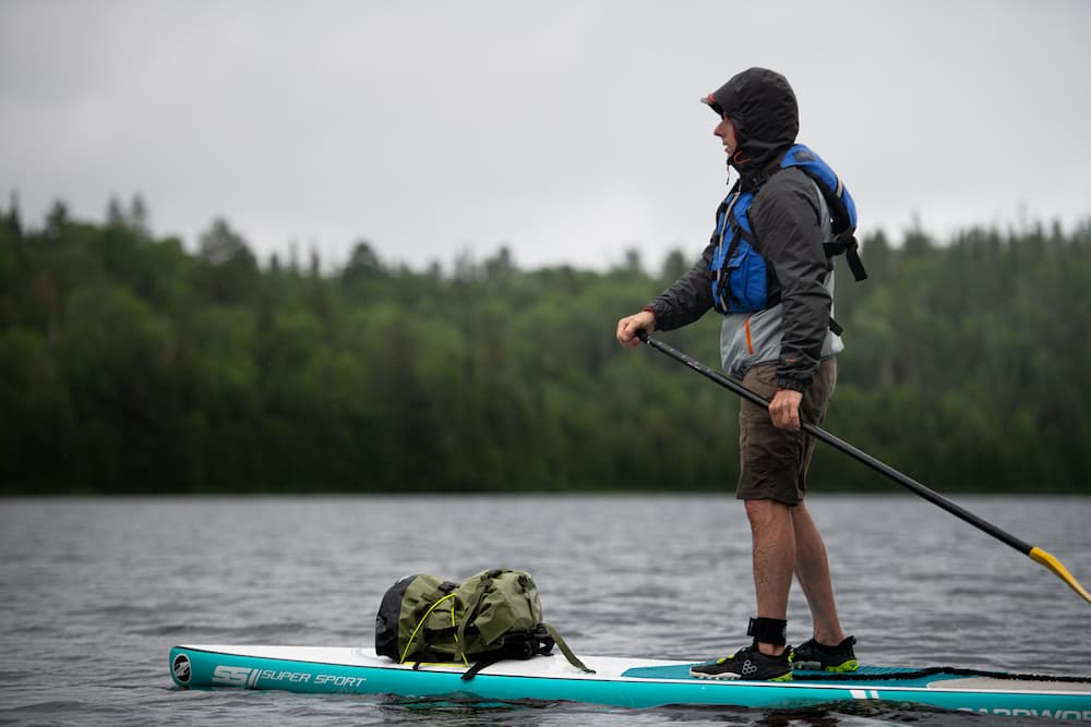 A person stands on a paddleboard on a lake, holding a paddle. The board is teal and white with a 55L idrybag expedition submersible green waterproof backpack resting on it.