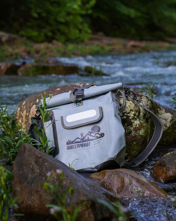 A idrybag light gray waterproof pro shield roll-top bag with a black strap and logo, resting on large rocks in a flowing stream with a forest background.