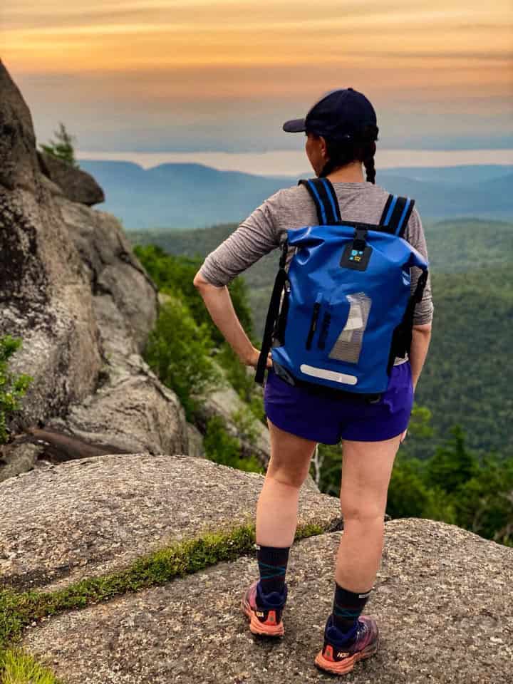 A woman stands on a rocky mountain peak at sunset wearing a blue idrybag pro lite waterproof backpack, purple shorts, and hiking shoes, with a scenic mountain landscape in the background.
