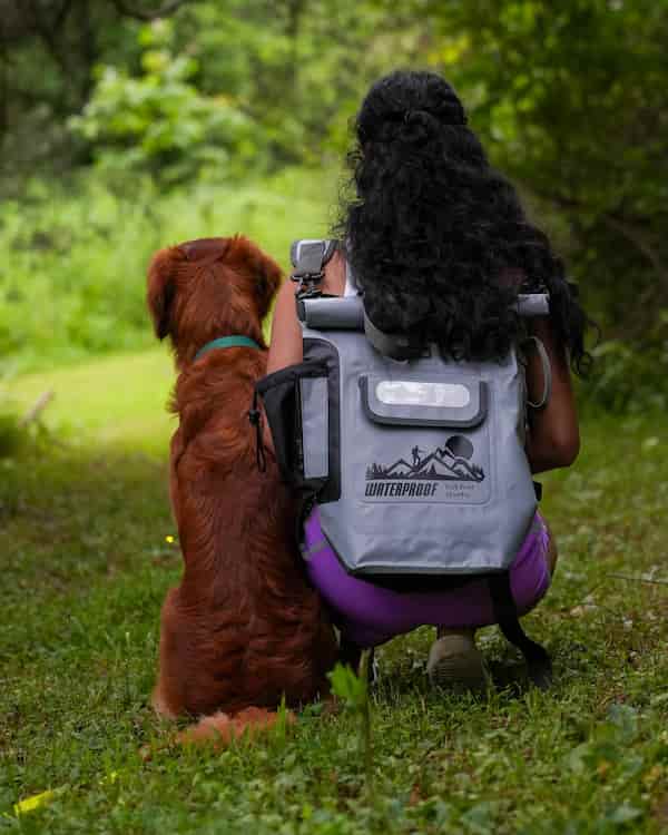 A woman crouches outdoors on grass next to a golden retriever dog, wearing a large idrybag pro shield gray waterproof backpack with a mounted camera on top. The dog sits beside her looking at the camera.