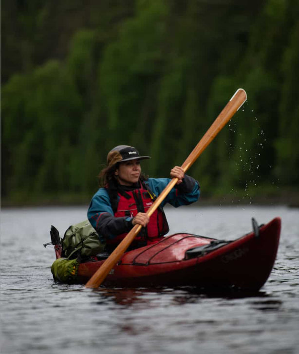 A person paddling a red canoe on a lake, holding a wooden paddle, with a forest background and water droplets visible. Having the 55L idrybag expedition submersible backpack