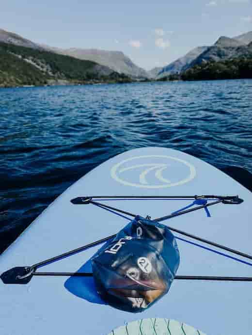 A blue inflatable stand-up paddle board on blue water with mountains in the background. A black 2L idrybag essential mate dry bag is strapped to the front of the board, and part of a paddle is visible.